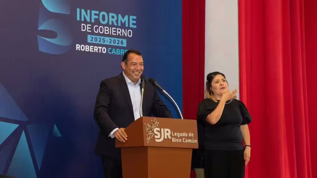 hombre-de-traje-sonrie-frente-a-un-podio-junto-a-una-interprete-de-lengua-de-senas-con-la-lona-del-quinto-informe-de-gobierno-a