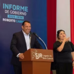 hombre-de-traje-sonrie-frente-a-un-podio-junto-a-una-interprete-de-lengua-de-senas-con-la-lona-del-quinto-informe-de-gobierno-a